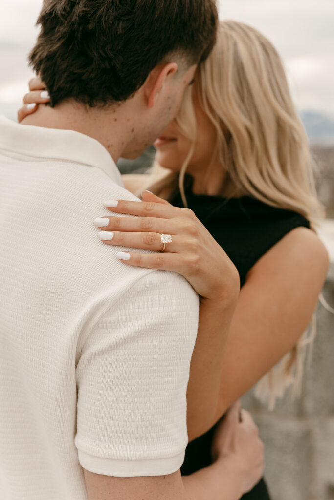 Close-up of engagement ring resting on fiancé’s shoulder during soft embrace