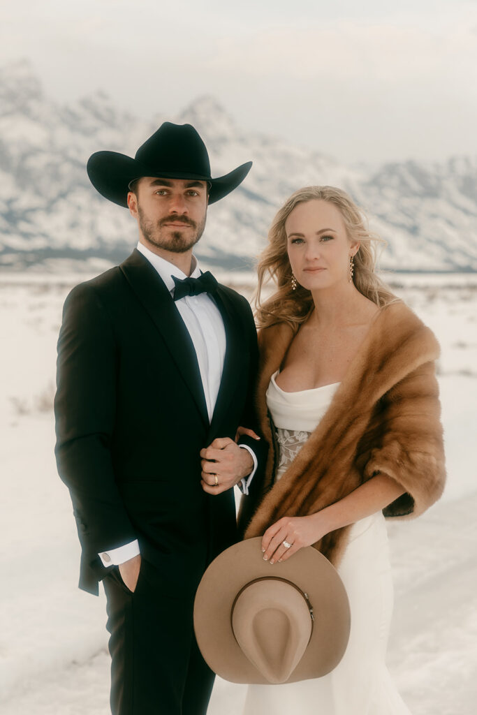Couple poses in fur and cowboy hat, framed by winter scenery and mountain views