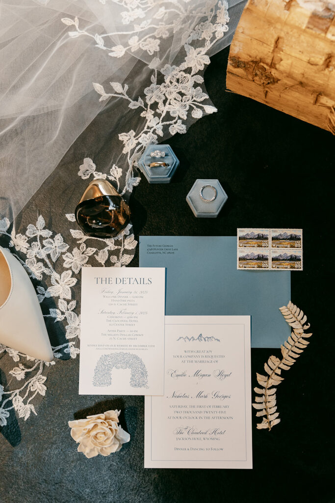Invitation suite flat lay with veil, rings, perfume, stamps, and floral accents