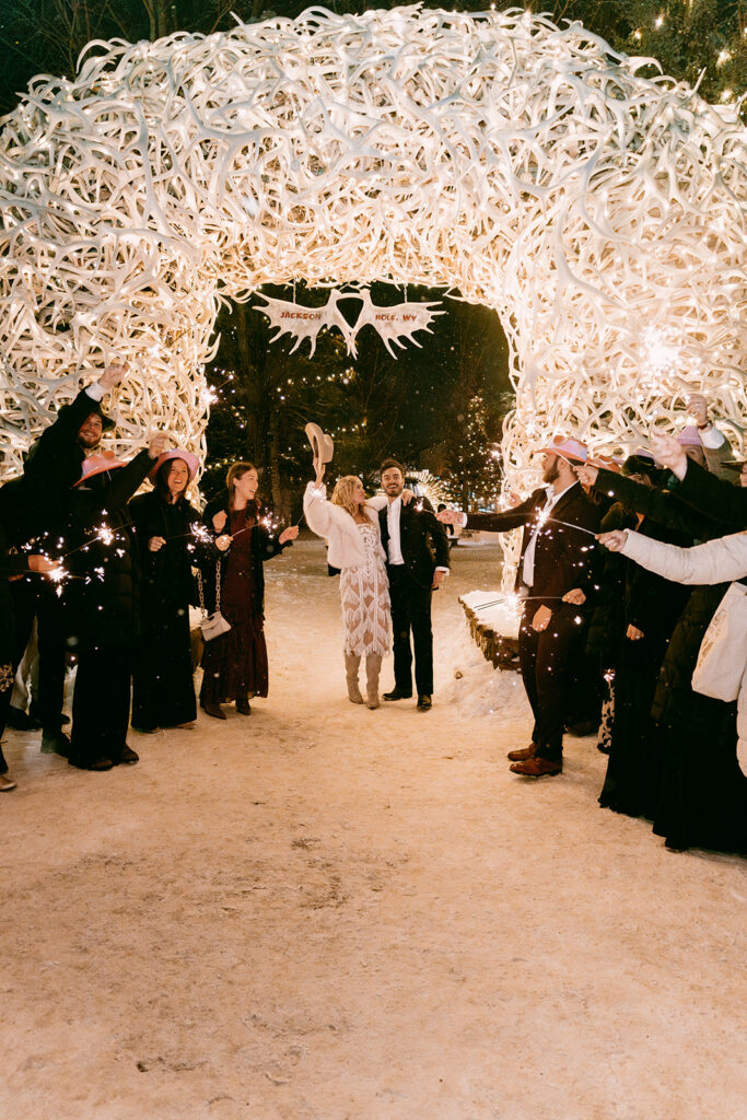 Couple exits under antler arch with sparklers and friends celebrating in winter evening light