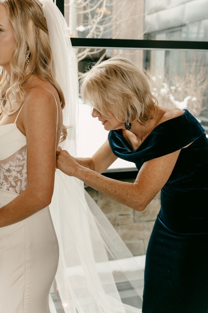 Bride’s mother helps button her dress as light streams in through large windows