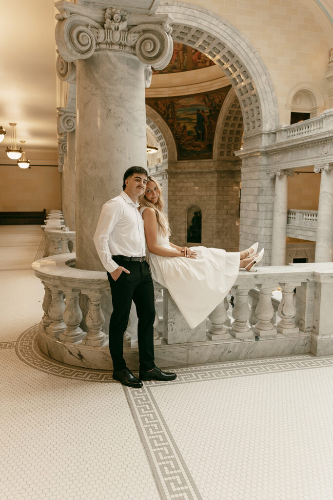 Bride sitting on marble ledge as groom stands beside her in Capitol interior