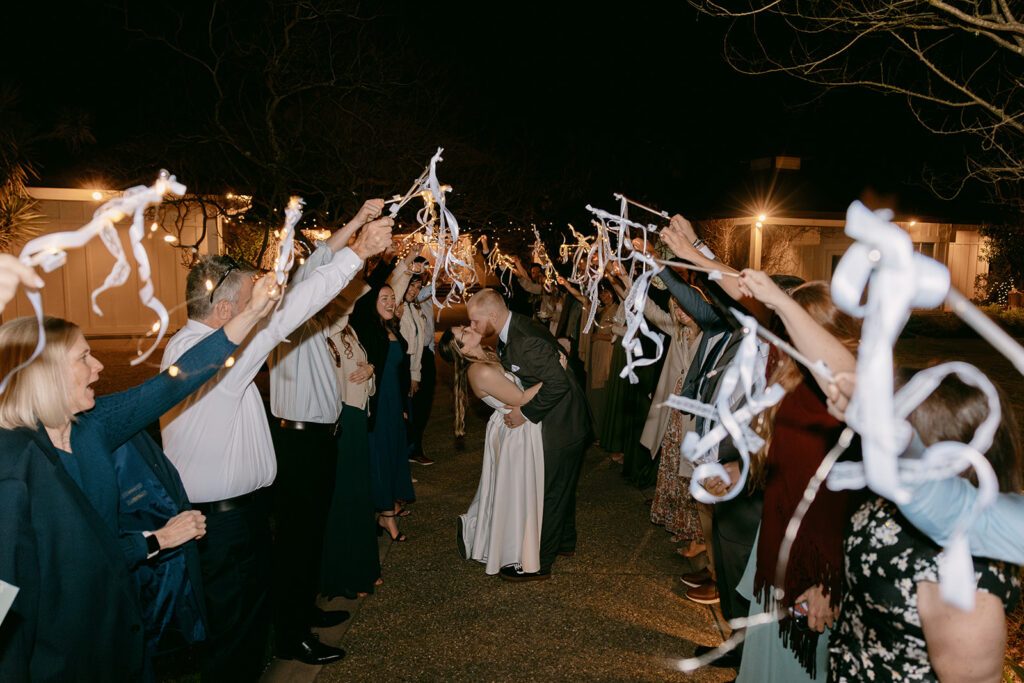 Couple shares a dip kiss during a nighttime ribbon wand send-off surrounded by cheering guests and twinkle lights
