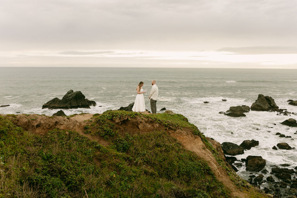 Bride and groom stand on a cliff above crashing waves, exchanging vows during their pre-wedding coastal session