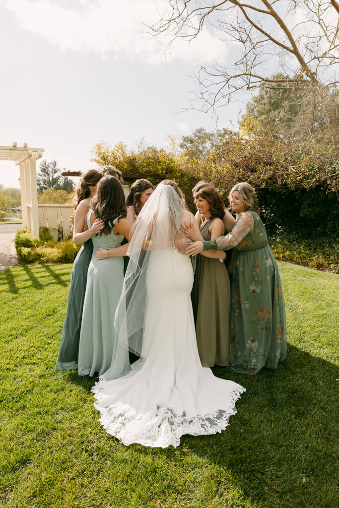 Bride surrounded by bridesmaids in green-toned dresses in a celebratory pre-ceremony hug.