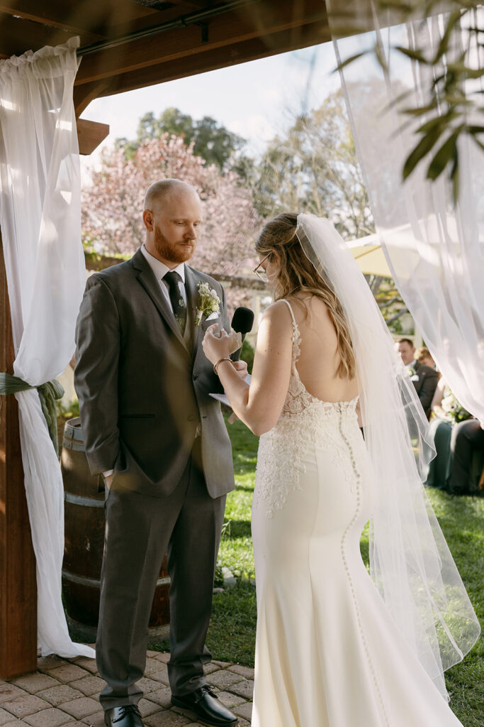 Bride reads vows to groom under a wooden arbor framed with flowing white drapes.