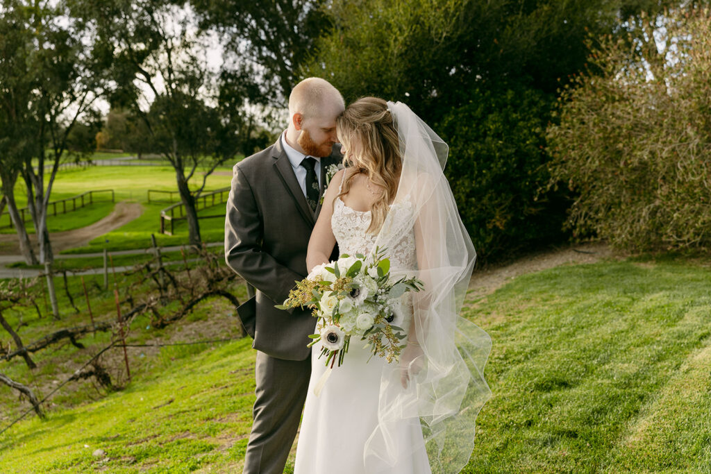 Bride and groom stand in the vineyard, forehead to forehead, holding a white bouquet.