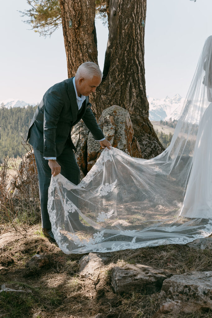 Father of the bride adjusts her veil near the ceremony site in the mountains.