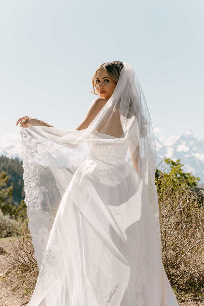 Bride looks over her shoulder, veil flowing in the breeze with alpine views.