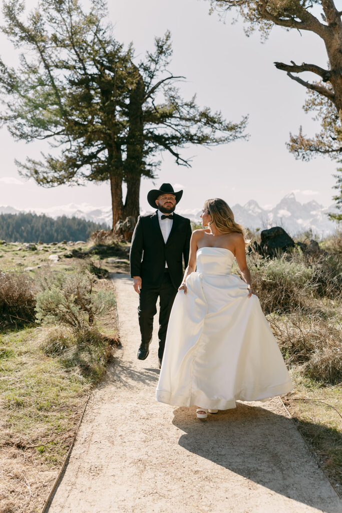 Bride and groom walk a tree-lined path, smiling at one another in the light.
