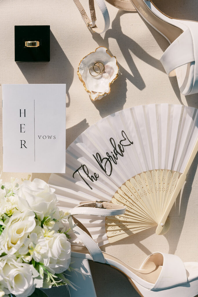 Flat lay of bridal details including vow book, fan, bouquet, heels, and rings in a shell dish.