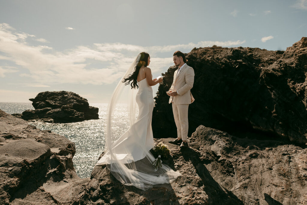 Couple exchanges handwritten vows on rocky cliffs with sparkling ocean behind them