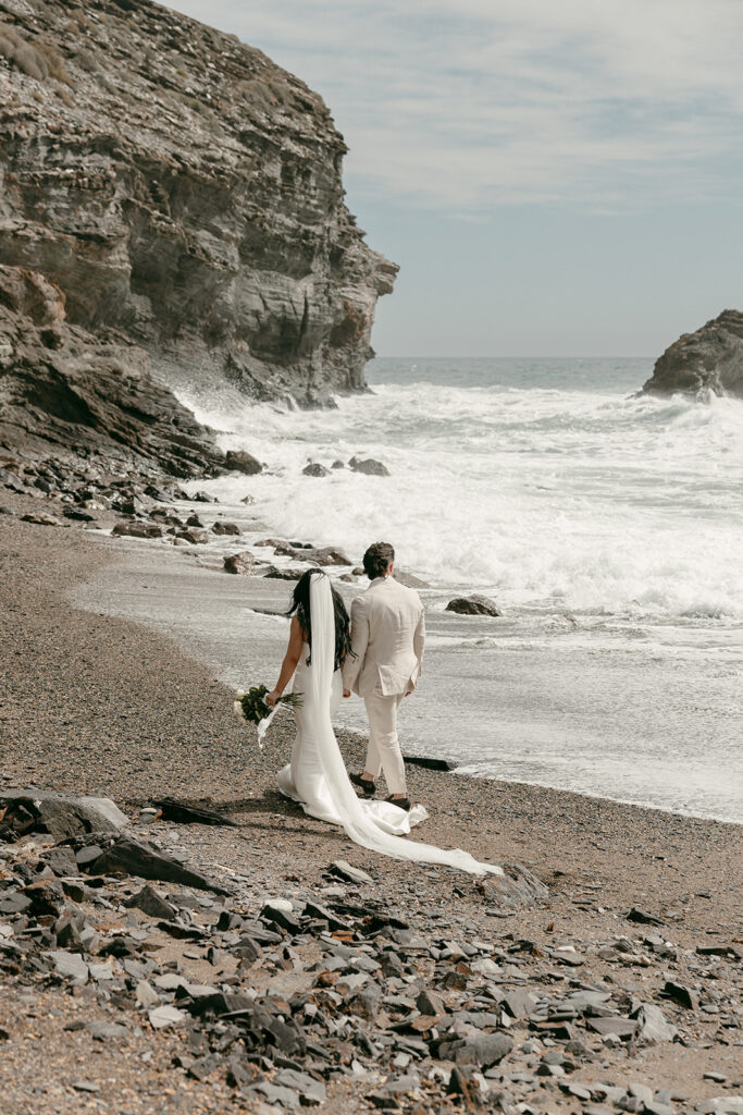 Bride and groom walk hand in hand along a secluded beach with crashing waves