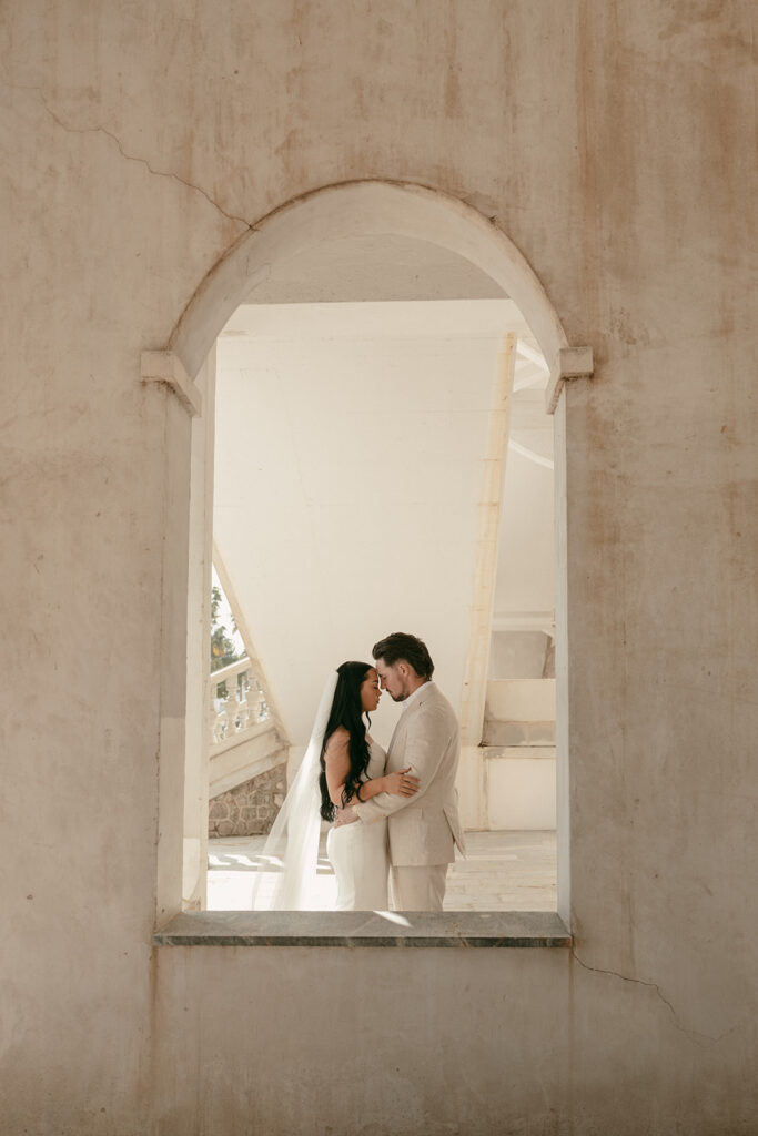 Bride and groom embrace in soft light framed by arched Spanish stone window