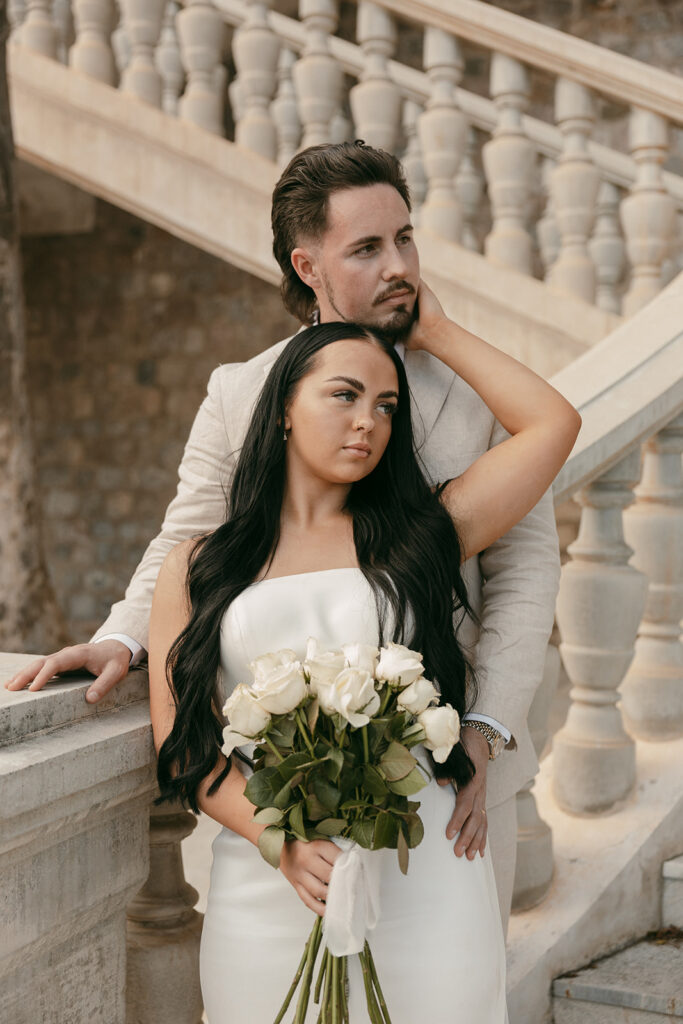 Bride holds bouquet while groom stands behind her on elegant stone staircase