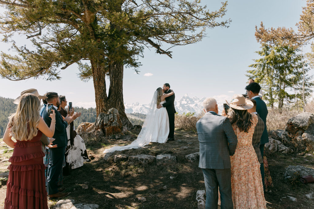 Guests witnessing the couple's first kiss beneath a large pine tree with the Tetons in the distance.