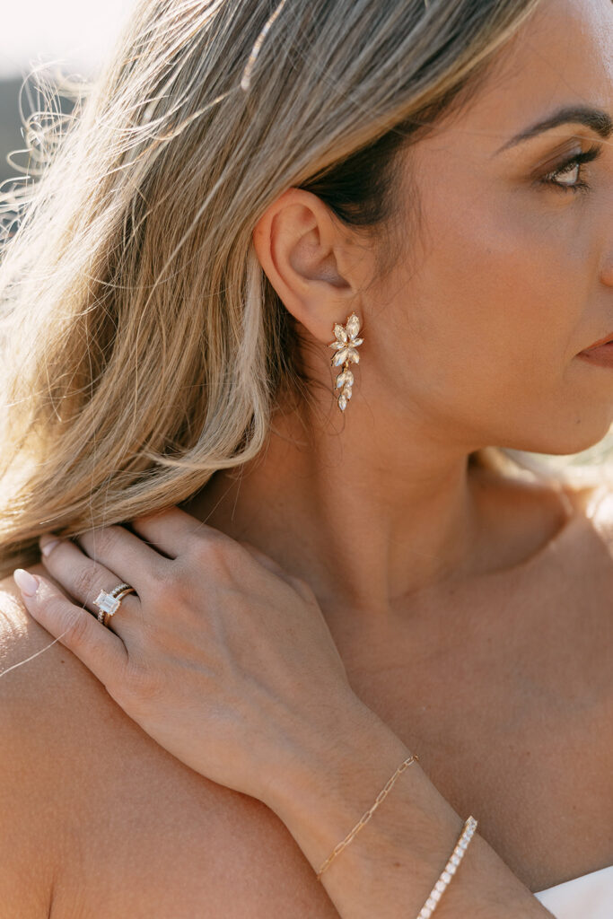 Close-up of bride’s gold floral earrings, engagement ring, and delicate bracelets.