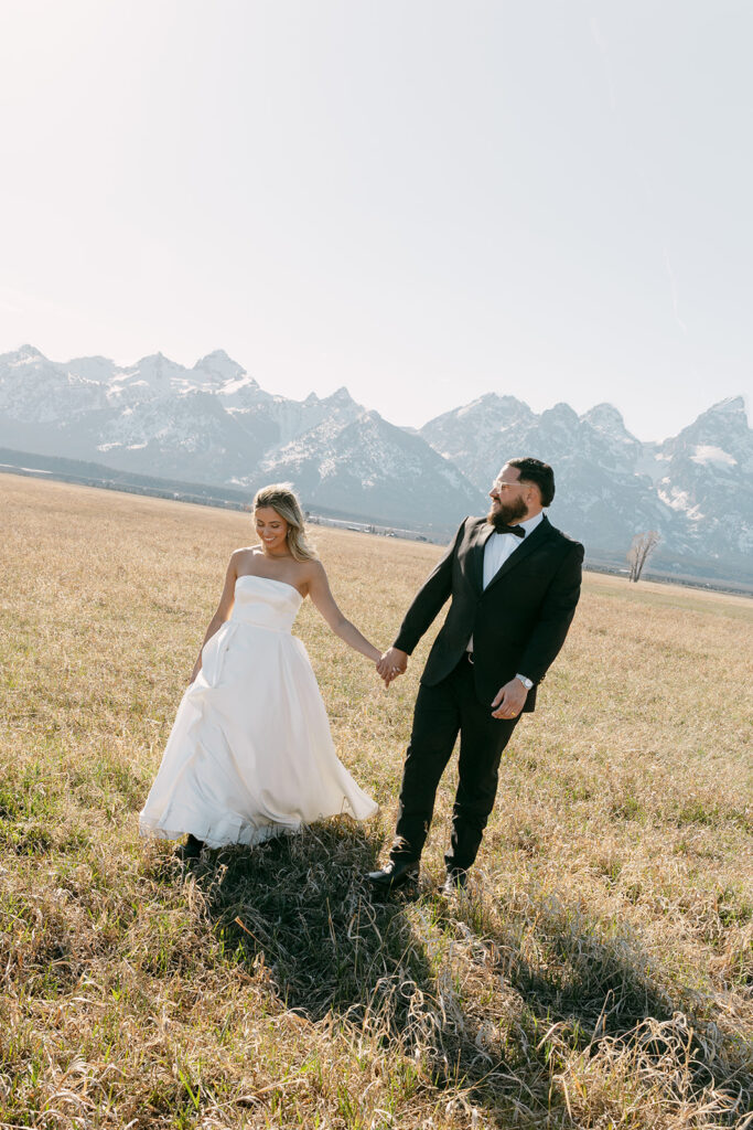 Bride and groom holding hands, walking through a field with Grand Teton peaks behind them.