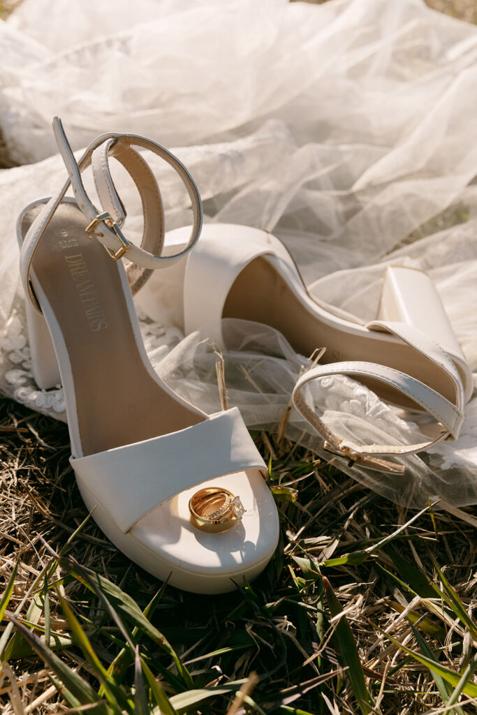 Wedding rings resting on a white heel with soft tulle and grass in golden hour light.