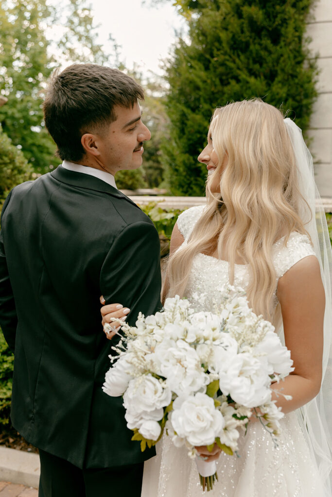 Bride and groom look at each other while holding bouquet of white florals during La Caille bridal portraits