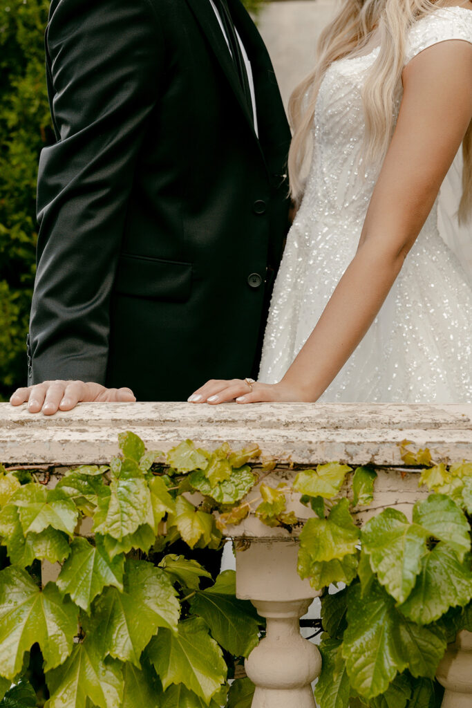Close-up of couple’s hands resting on ivy-draped railing, showing her engagement ring