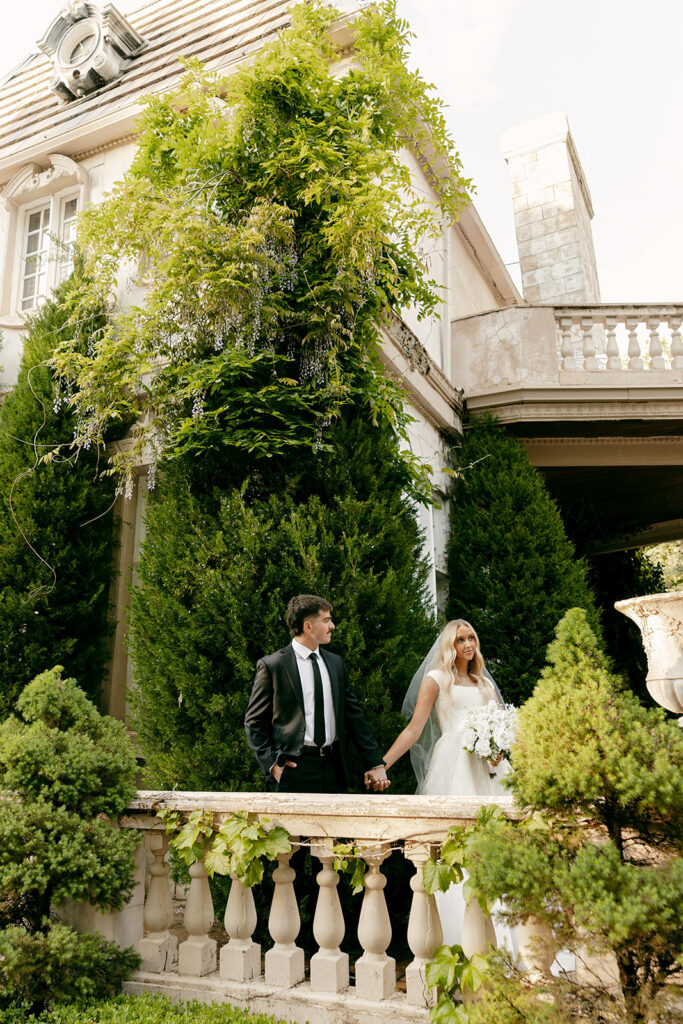 Bride and groom holding hands on a stone balcony framed by greenery