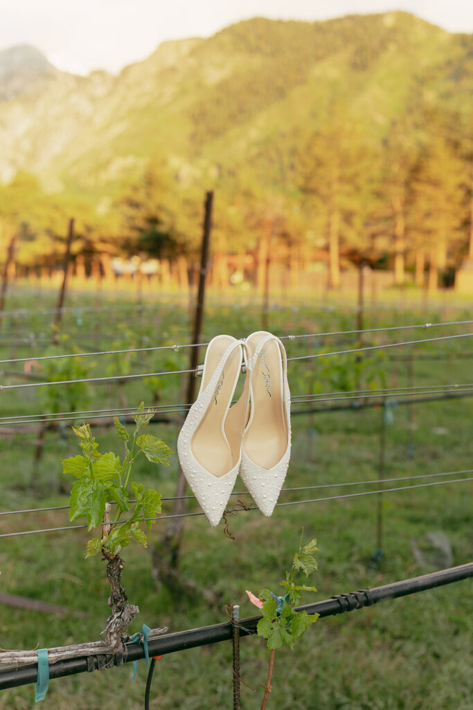 Bride’s pearl-embellished heels hang on a vineyard wire at sunset