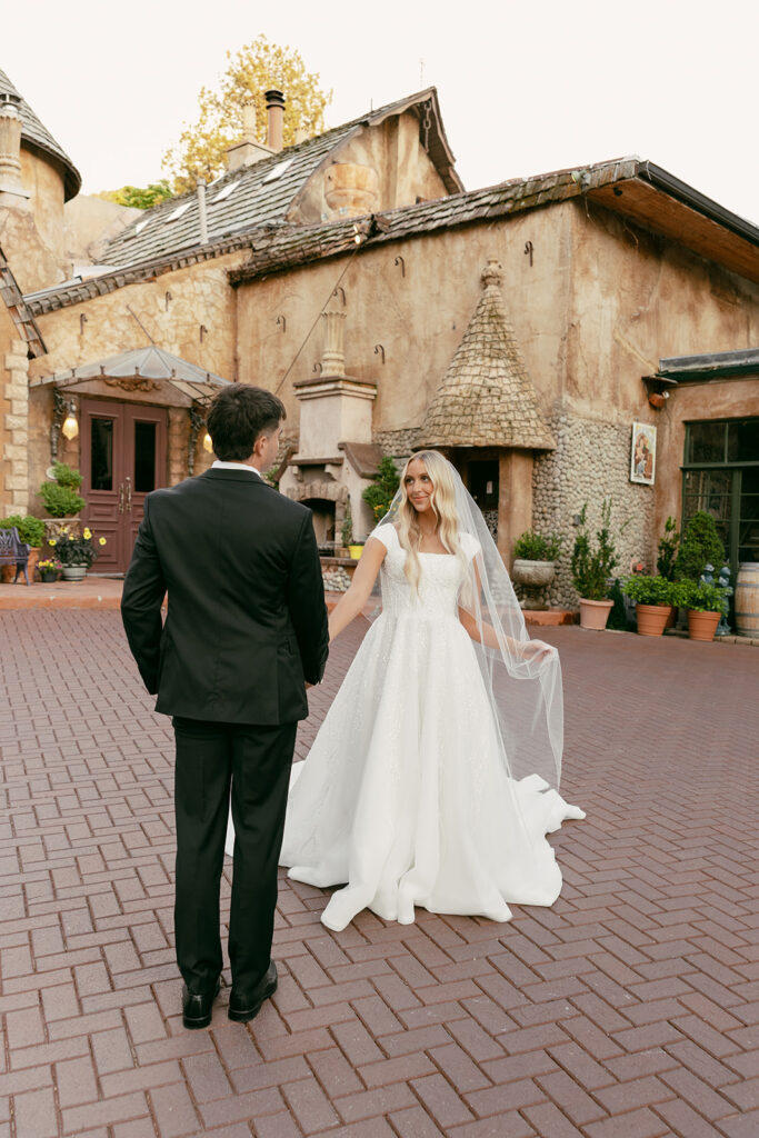 Bride and groom hold hands in front of a rustic stucco villa with turreted rooflines