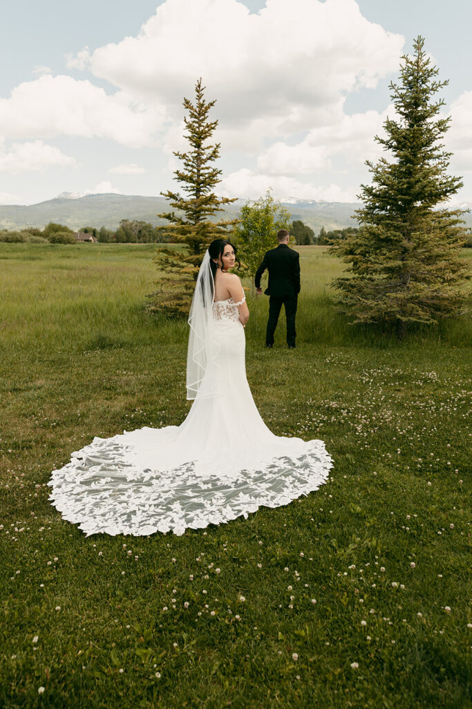 Bride prepares for first look, standing behind groom in an open field