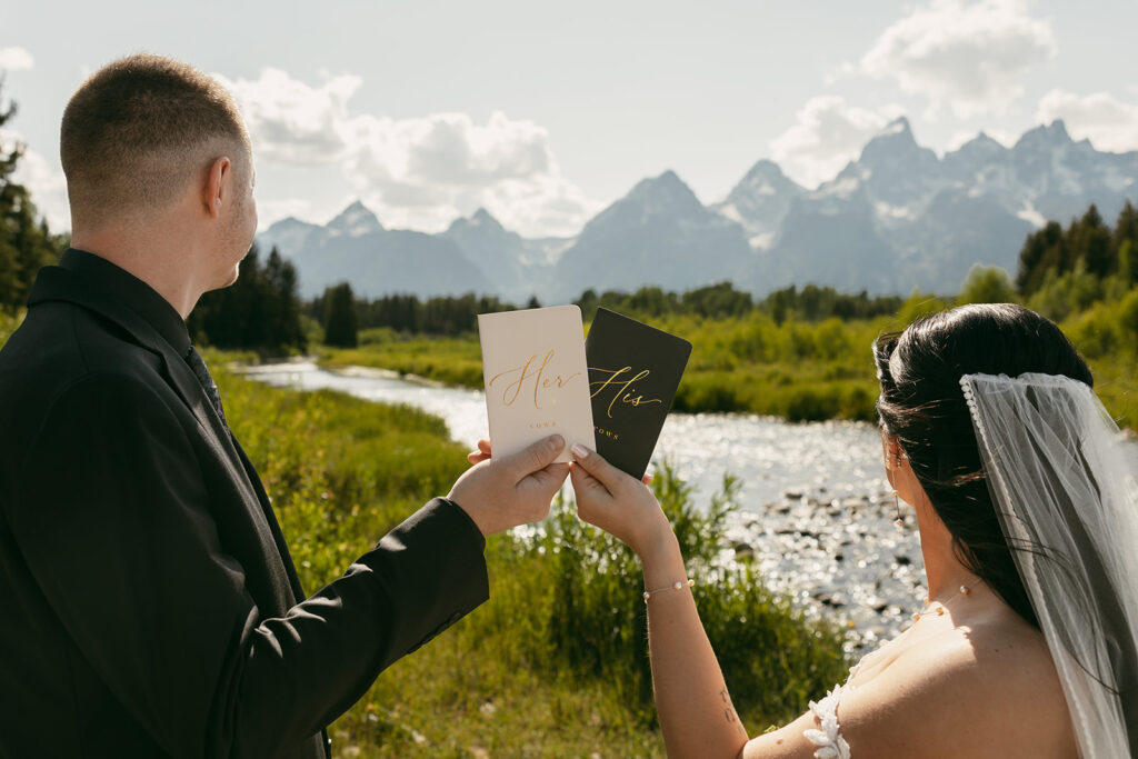 Bride and groom holding up vow books with Teton mountains and Snake River behind