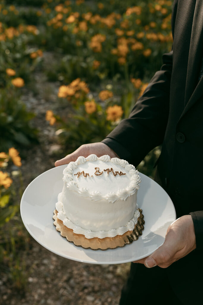 Groom hold Jackson Hole elopement cake in front of wildflowers