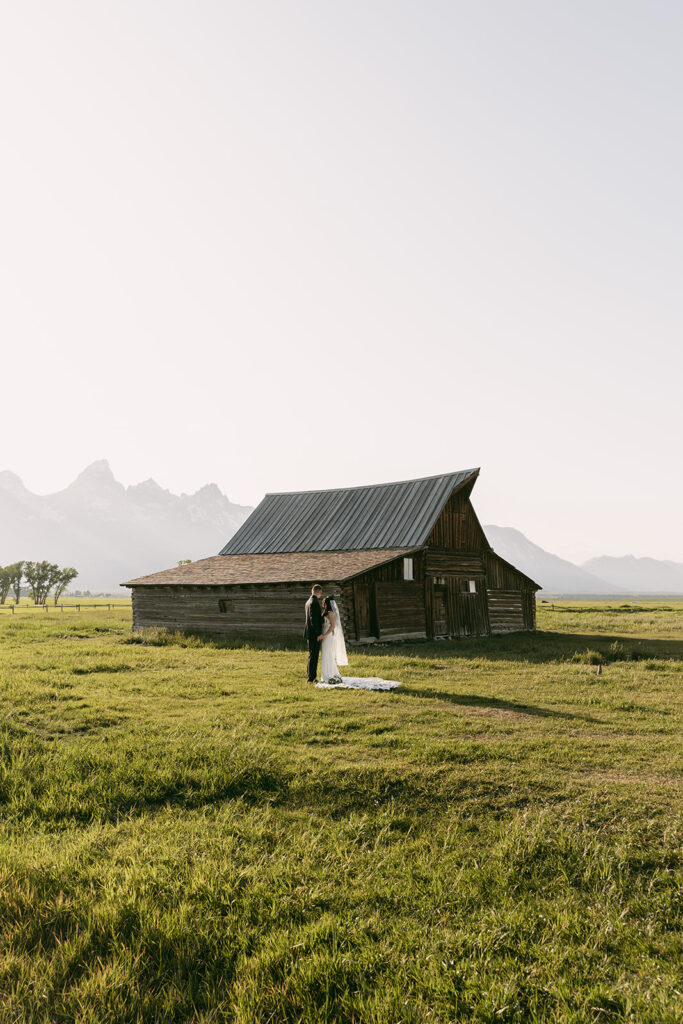 Wide shot of bride and groom standing in an open field with barn in background
