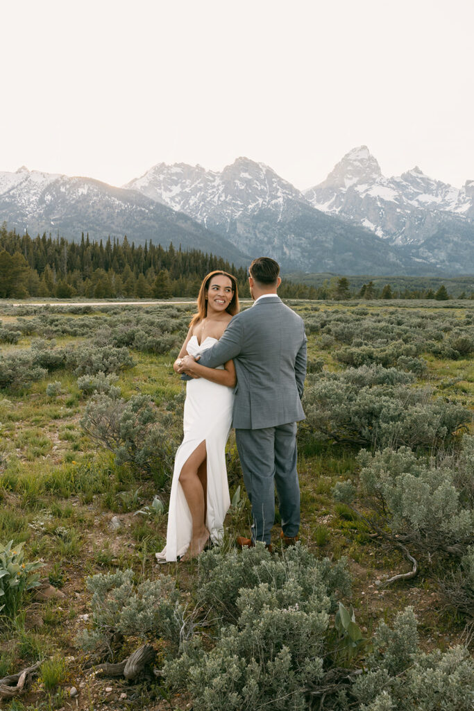 Bride smiles into camera as groom looks back at the Grand Teton mountain range