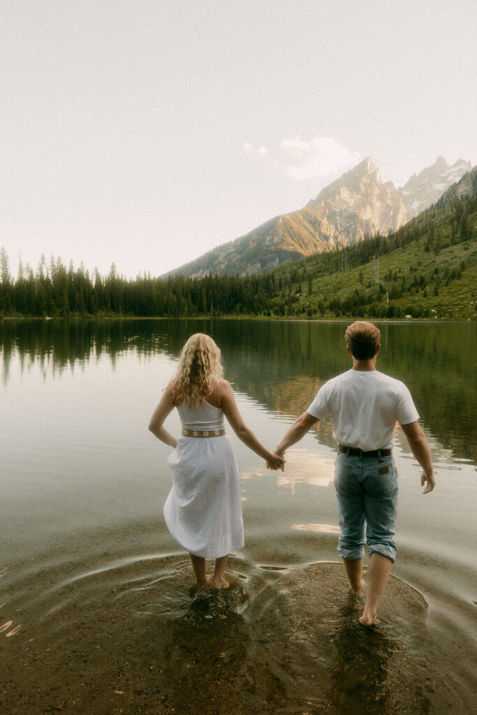Engaged couple wades out into String Lake with mountains in the distance