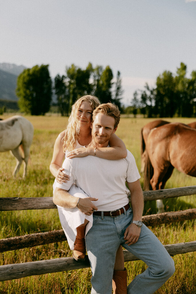 Couple poses on rustic fence with horses during romantic Tetons engagement photography session
