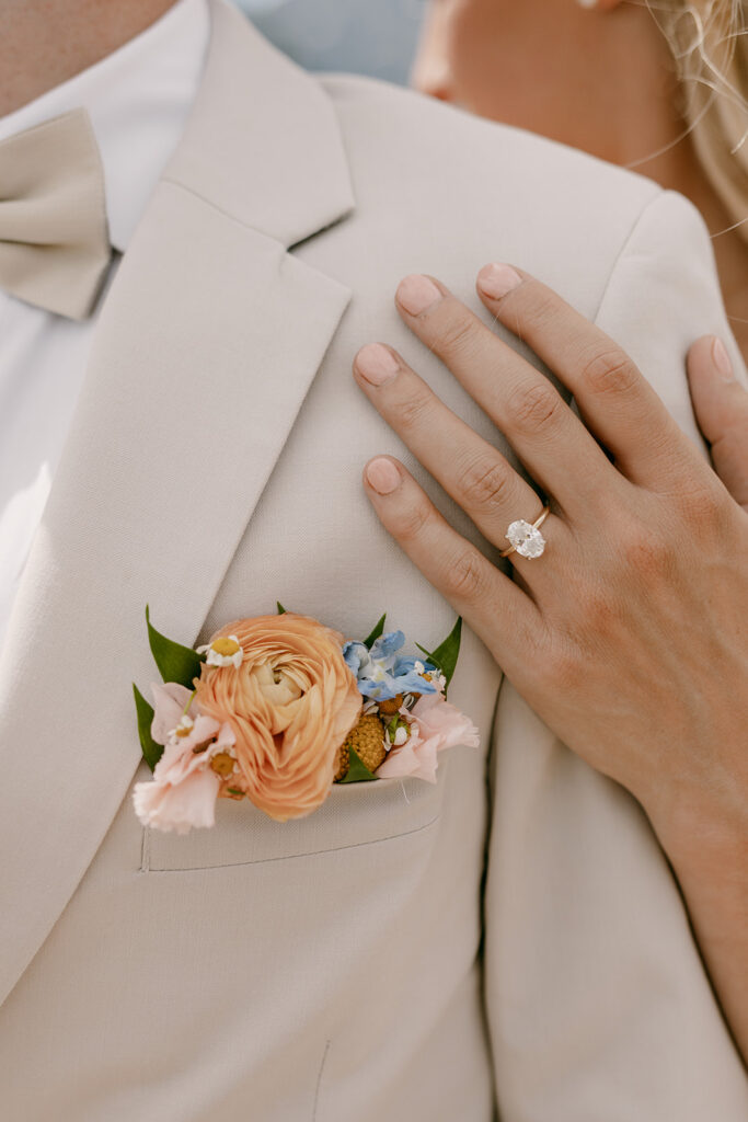 Close-up of vibrant orange, pink, and blue boutonniere and bride's ring hand
