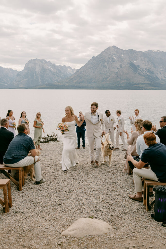 Bride and groom walk up the aisle with golden retriever and Grand Tetons in background