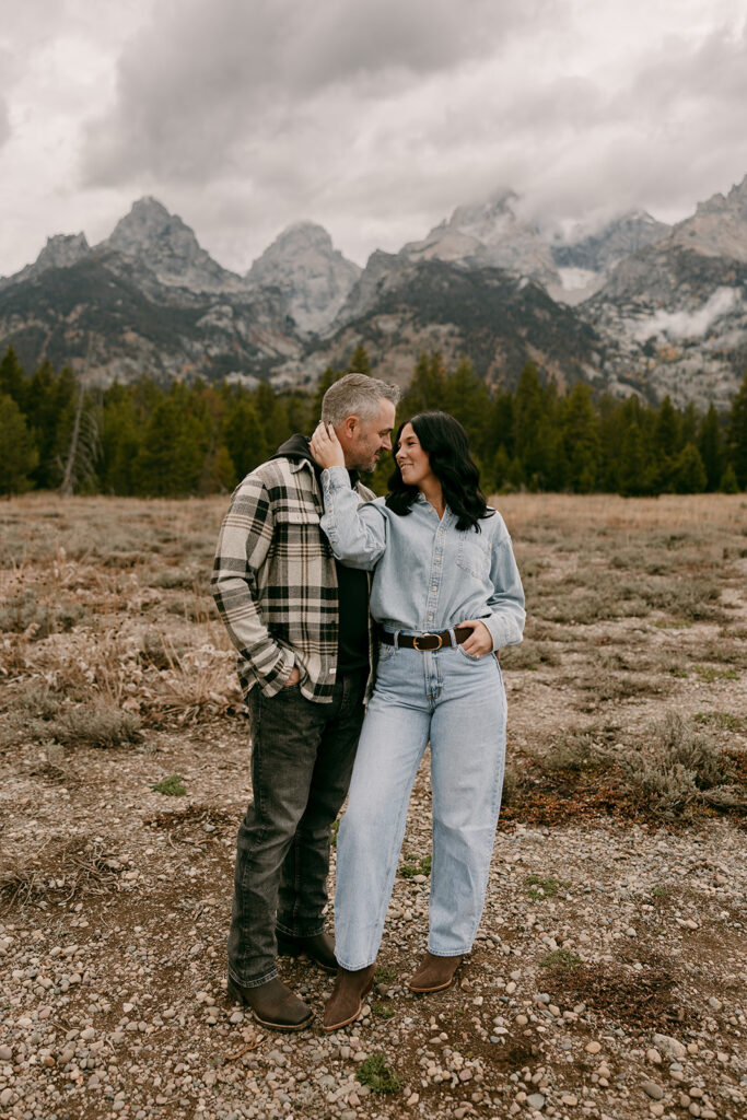 Couple standing forehead to forehead with the Grand Teton mountains rising behind them under a cloudy sky.