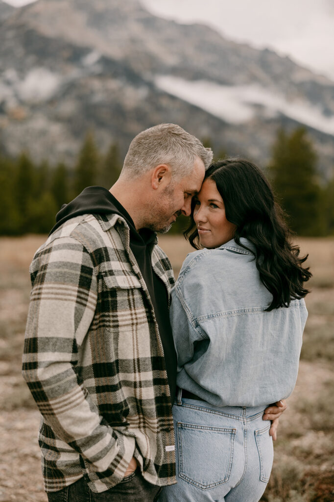 Romantic close-up of couple holding each other with mountain peaks and evergreen trees softly blurred in the background.