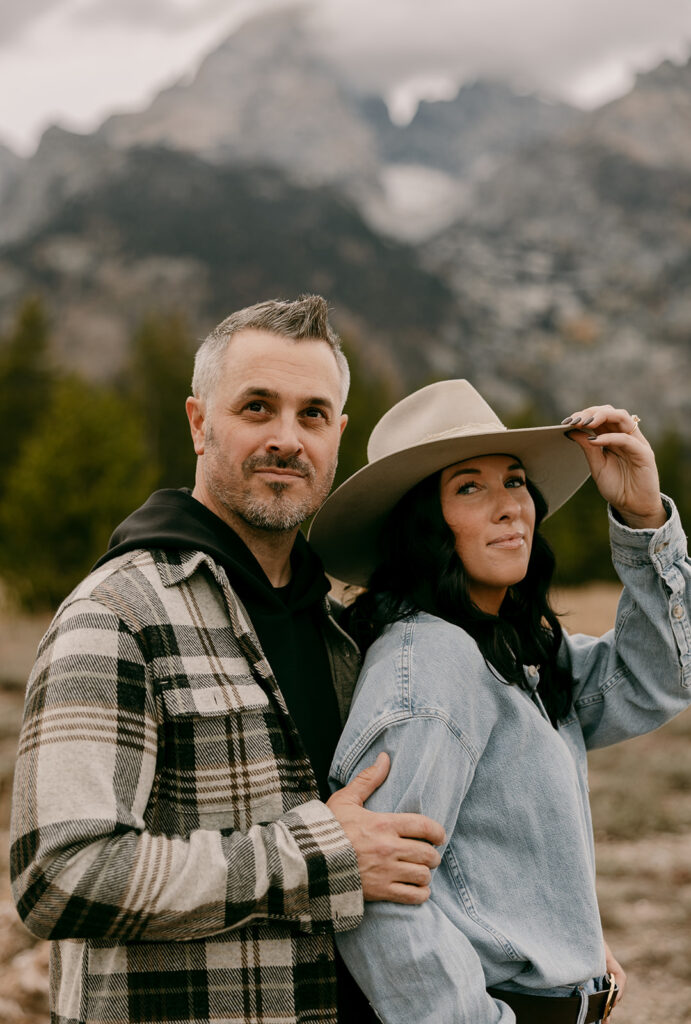 Couple poses confidently in denim and flannel with the Tetons behind them, the woman tipping her felt hat.