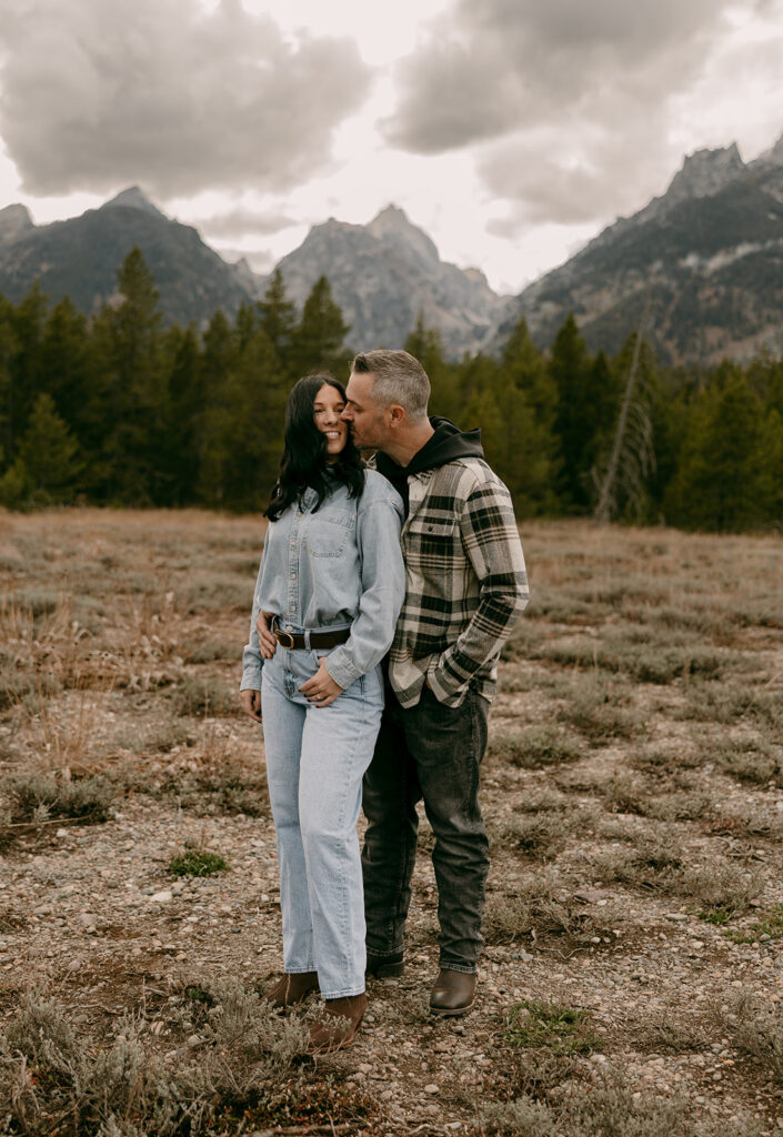 Smiling couple shares a candid kiss in front of a pine forest and rugged mountain skyline in Jackson Hole.