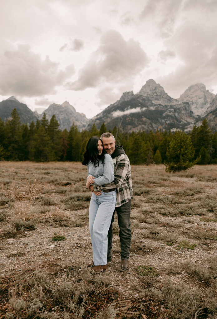 Man hugs his fiancée from behind as they laugh together in an open field during their Grand Tetons engagement photography session.