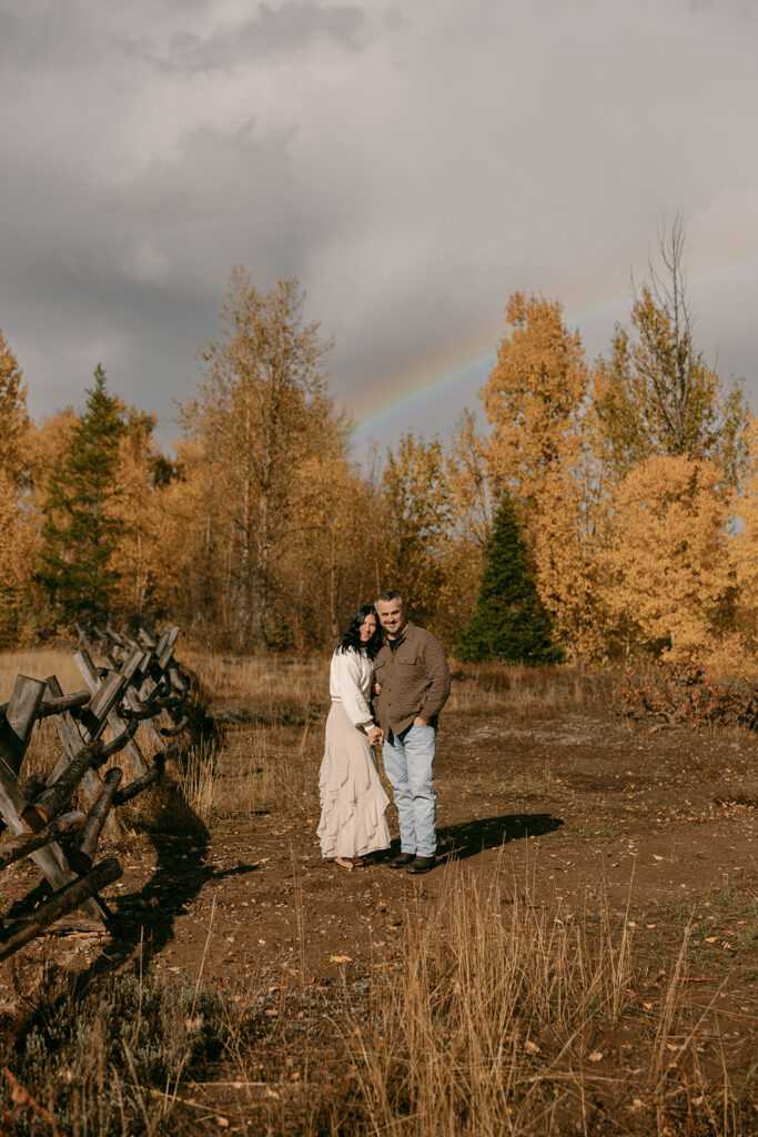 Couple embraces by a rustic fence under golden trees and a faint rainbow during a romantic autumn session.