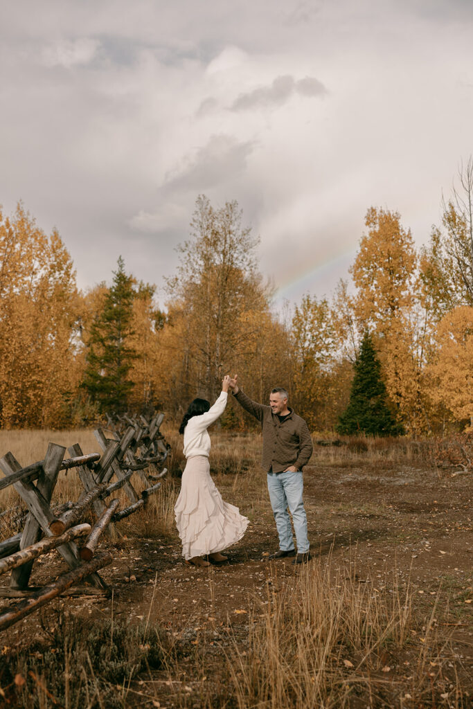 Joyful couple dances beneath a rainbow, framed by fall leaves and wooden fencing near Mormon Row.