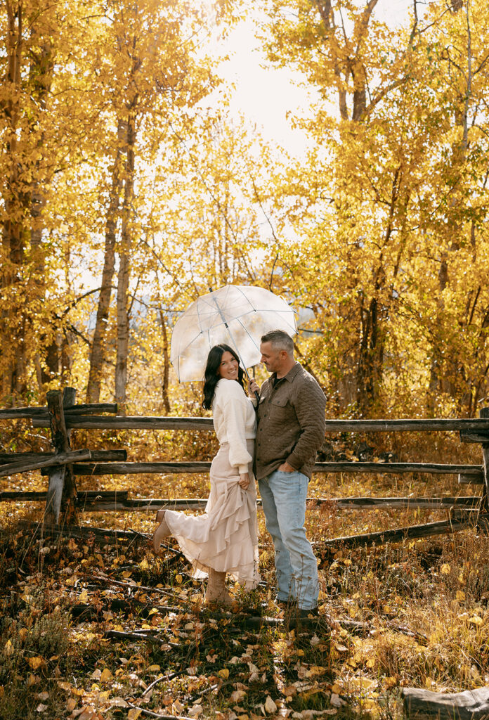 She smiles with one leg lifted, twirling under a clear umbrella beside her fiancé in a western-inspired outfit.