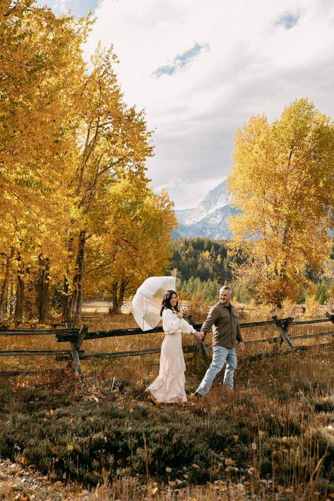 Walking hand in hand with a clear umbrella, the couple crosses a leaf-lined path in Grand Teton National Park.