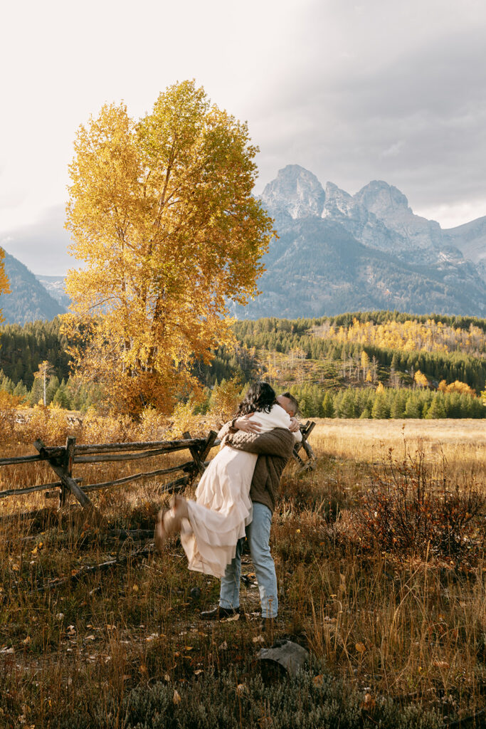 Swept into his arms, she twirls with laughter at the base of the mountain peaks during Grand Tetons engagement photography.