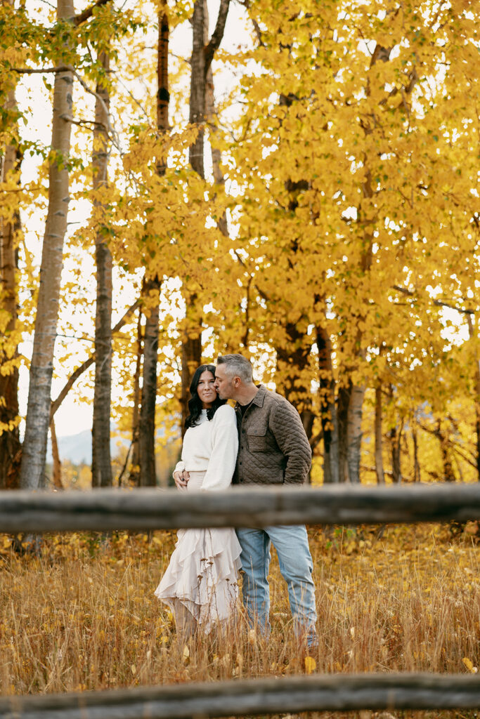 Man kisses woman’s forehead as they stand in front of a rustic fence beneath golden fall foliage.