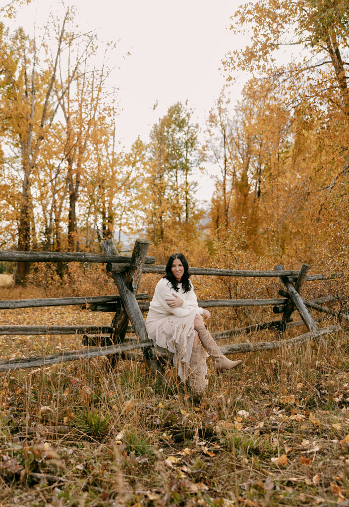 Woman in boots and sweater sits on a weathered wood fence surrounded by golden fall trees.