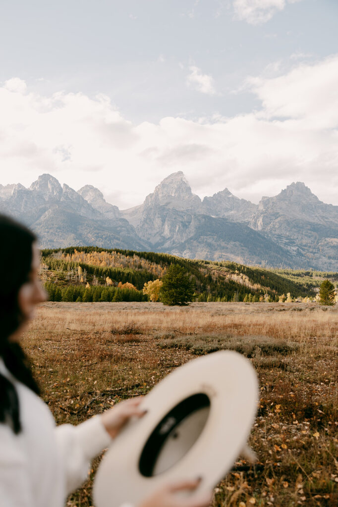 Woman holds wide-brim hat while admiring the Grand Teton mountain range across a golden valley.